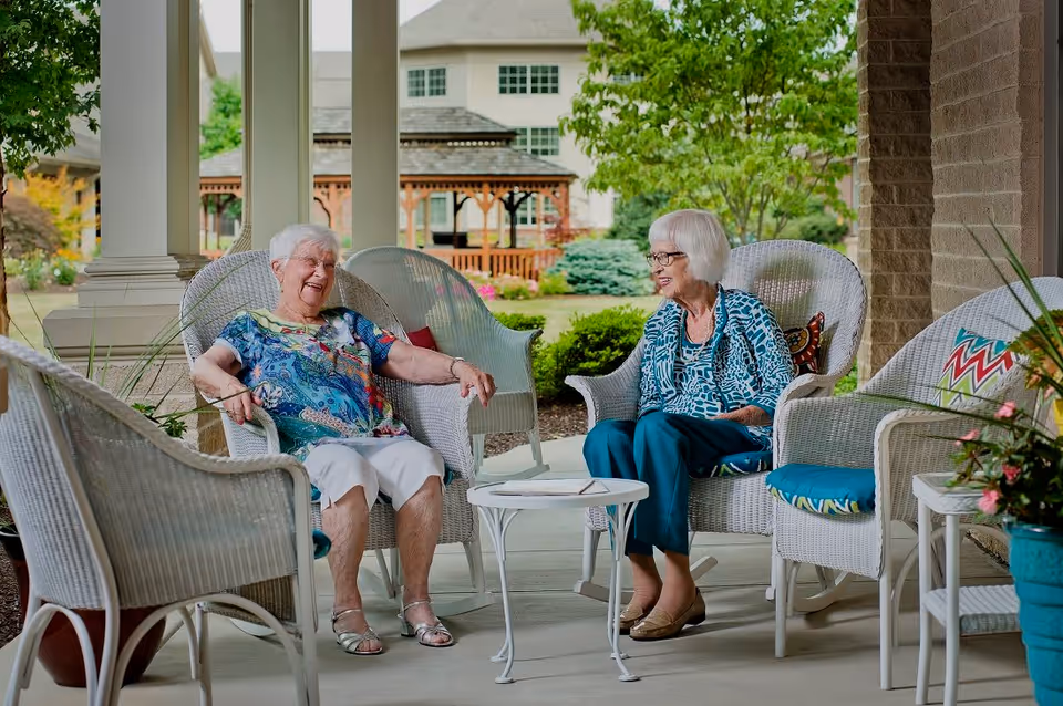 Two elderly women sitting and chatting on white wicker chairs on a covered patio with a small white table between them. The background shows a garden with trees, shrubs, and a wooden gazebo.