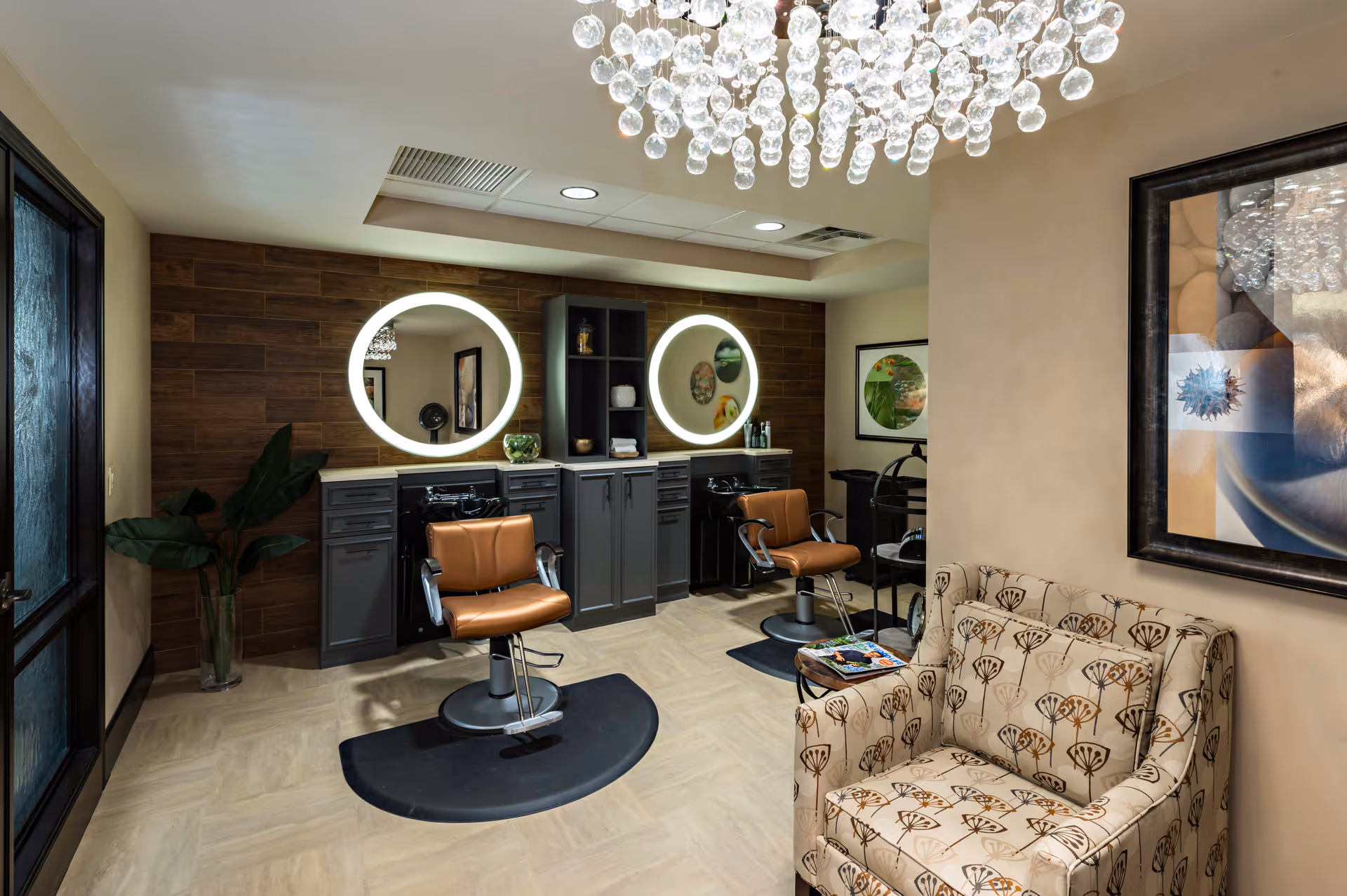 Interior view of a stylish salon area with two brown salon chairs in front of illuminated round mirrors mounted on a wooden accent wall. There is a patterned armchair and a small table with magazines in the foreground, a decorative plant in a glass vase to the left, and framed artwork on the walls. A modern chandelier with hanging glass orbs is visible on the ceiling.