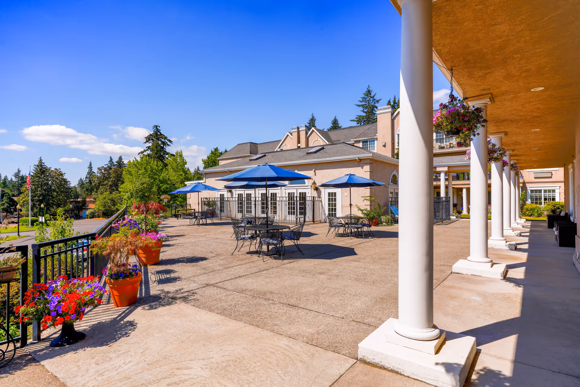 Outdoor patio area at a senior living facility with several round tables and chairs under blue umbrellas, potted flowers along the railing, white columns supporting an overhead structure, and a building in the background under a clear blue sky.