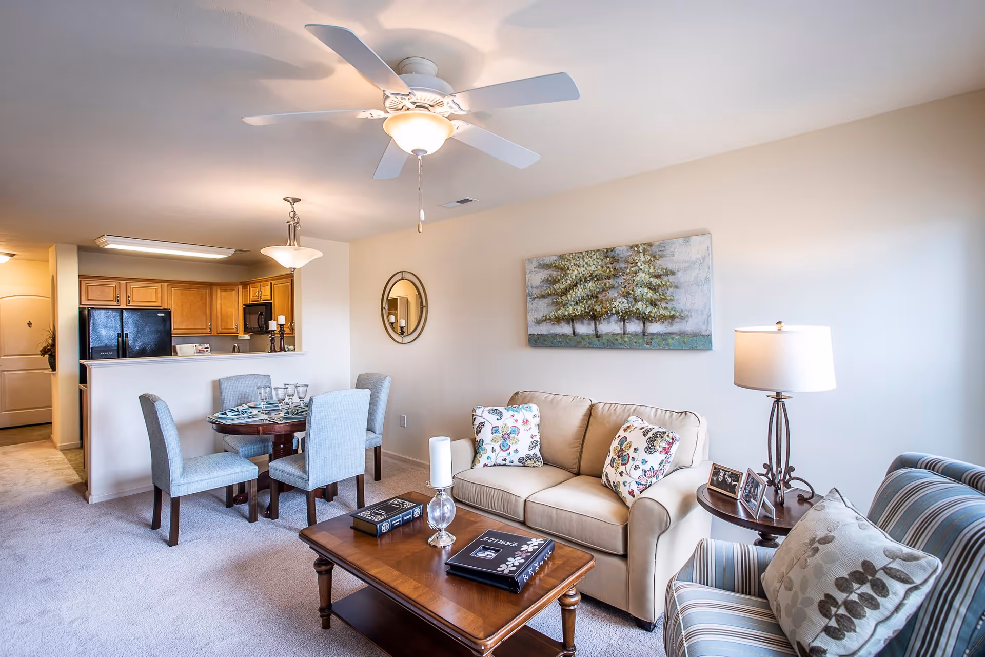 A bright and cozy living room and dining area in an apartment. The living room features a beige sofa with floral pillows, a striped armchair with a decorative pillow, a wooden coffee table with books and candles, and a side table with a lamp and framed photos. The dining area has a round table set with four upholstered chairs and place settings. The kitchen with wooden cabinets and black appliances is visible in the background. A ceiling fan with a light fixture is mounted on the ceiling, and a painting of trees hangs on the wall above the sofa.