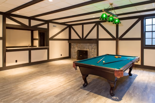 A spacious game room with a pool table in the center, featuring a green felt surface and pool cues resting on it. The room has wood-patterned flooring, beige walls with dark wood trim, a stone fireplace, and a window letting in natural light. A green hanging light fixture is above the pool table.