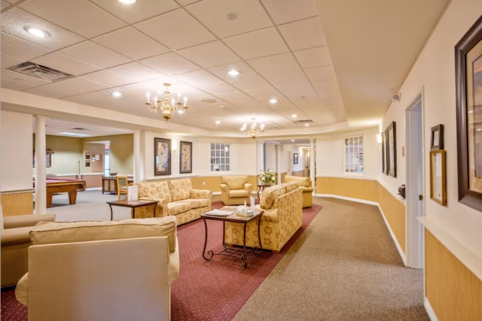A spacious and well-lit common area in a senior living facility featuring multiple beige and floral-patterned sofas arranged around small tables. The room has a carpeted floor with a red rug in the seating area, white walls with wooden paneling, framed artwork, and chandeliers hanging from a drop ceiling. In the background, there is a pool table and additional seating visible through an open doorway.