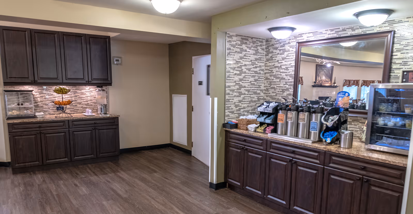 Interior view of a senior living community beverage and snack station area with dark wood cabinets, a large mirror, multiple coffee dispensers, a small refrigerator with drinks, and a counter with snacks and condiments. The floor is wood, and the walls have a beige and tiled backsplash design.