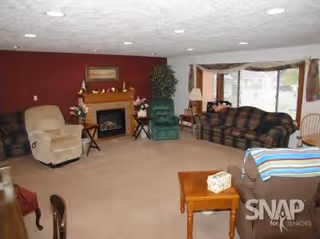A cozy living room with beige carpet, a fireplace with decorations on the mantle, a green recliner, a beige recliner, a plaid sofa, a wooden side table with a tissue box, and large windows with curtains letting in natural light.