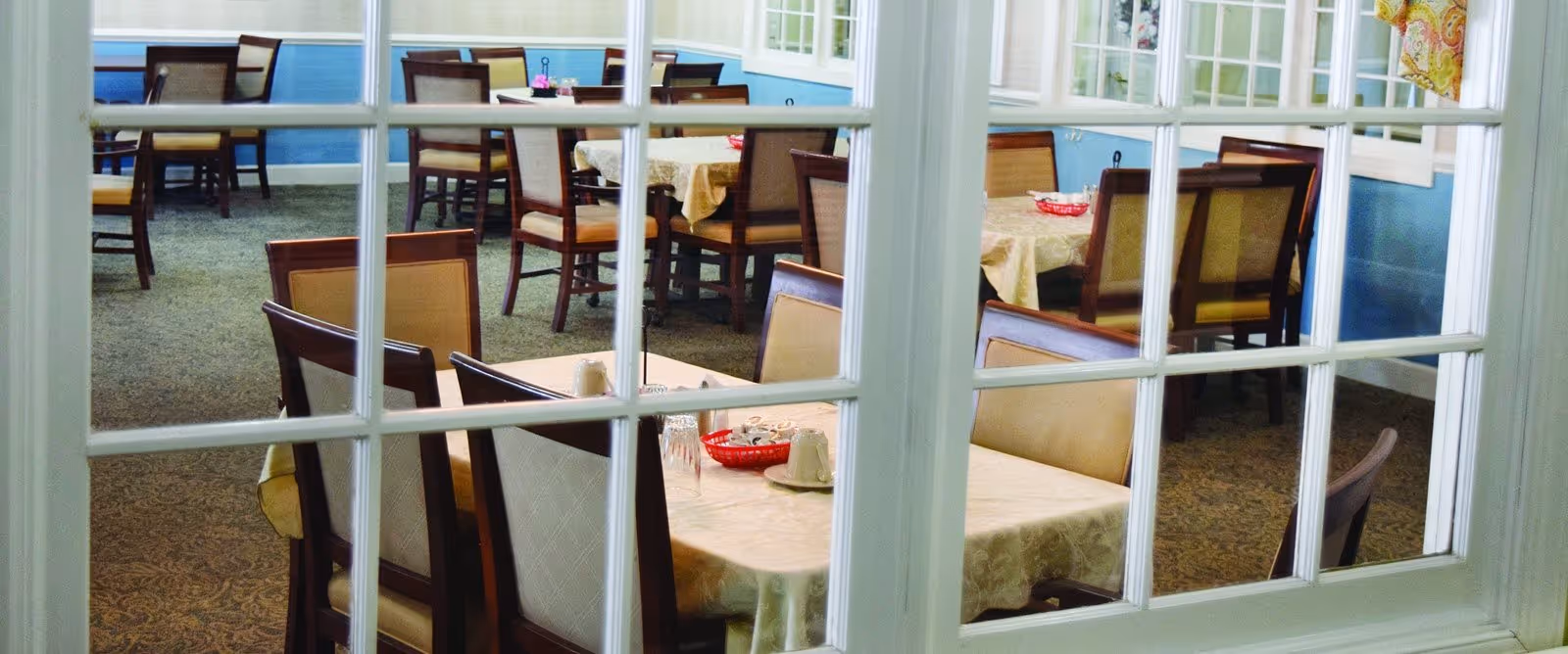 View through multi-pane glass into a dining room with several tables and chairs set with tablecloths.