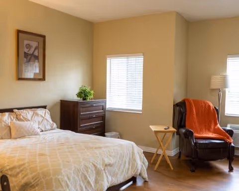 A cozy bedroom in a senior living facility featuring a bed with beige patterned bedding, a dark wooden dresser with a small green plant on top, a framed artwork on the wall, a window with white blinds, a small wooden side table, a dark leather armchair with an orange throw blanket, and a floor lamp next to the chair.
