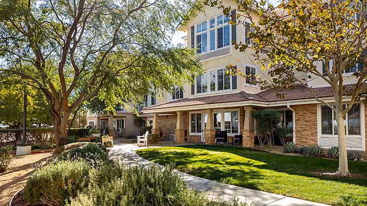 Outdoor view of Holiday Rancho Village showing a well-maintained garden with trees, shrubs, and a paved walkway leading to a multi-story building with large windows and a covered patio area with chairs.