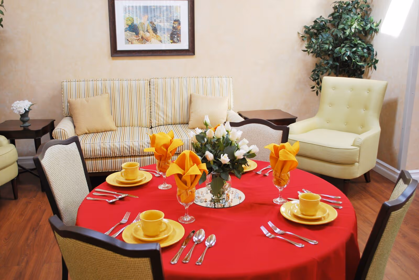 A cozy dining area with a round table covered in a red tablecloth, set with yellow plates, cups, and orange napkins in glasses. The table has a centerpiece of white flowers. Surrounding the table are cushioned chairs, a striped sofa with two beige pillows, a light green armchair, and a potted plant in the corner. A framed picture hangs on the beige wall behind the sofa.