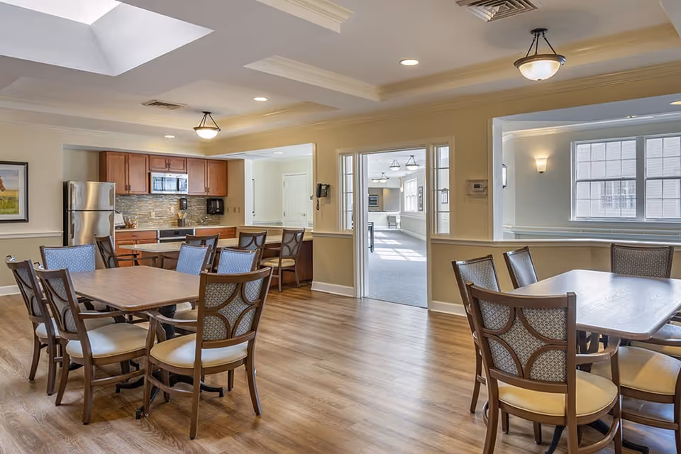 A bright and spacious dining area with wooden tables and cushioned chairs. The room features a kitchen area with wooden cabinets, a stainless steel refrigerator, microwave, and a tiled backsplash. The floor is wood, and there are large windows and ceiling lights providing ample natural and artificial light.