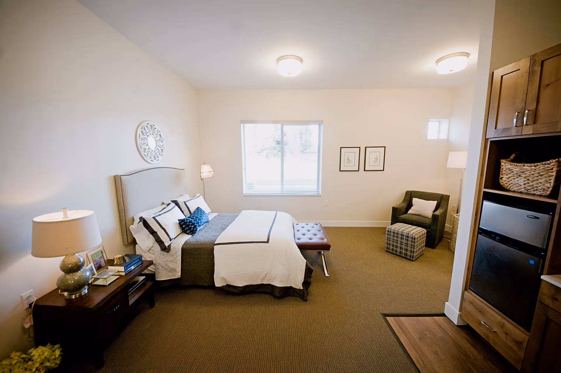 A well-lit bedroom in an assisted living facility featuring a neatly made bed with white and gray bedding, a bedside table with a lamp and books, a cushioned bench at the foot of the bed, a comfortable armchair with a pillow and a plaid ottoman, a window with blinds, framed artwork on the wall, and a built-in wooden cabinet with a mini refrigerator and basket.