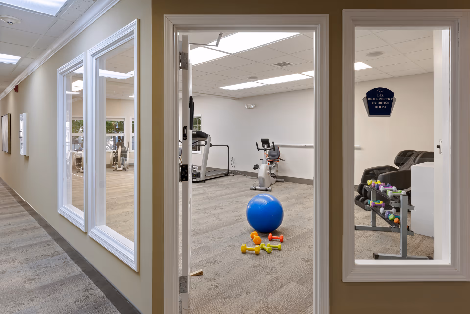 View through a hallway window and open door into a senior living facility exercise room containing a blue exercise ball, colorful dumbbells on the floor and on a rack, a stationary bike, and a treadmill. The room has beige walls and carpeted flooring with bright overhead lighting.