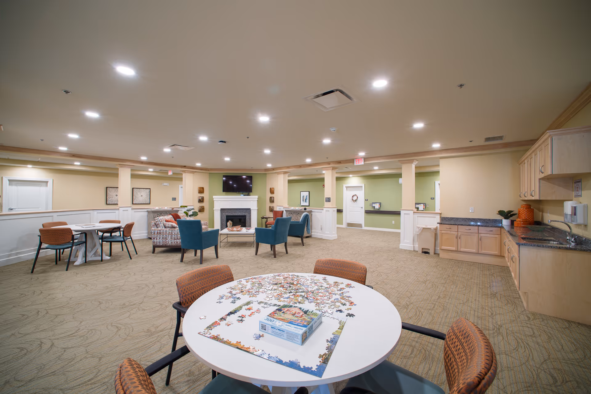 A spacious senior living common area with a round table in the foreground featuring a partially completed jigsaw puzzle. The room has beige carpet, light-colored walls with green accent walls, recessed ceiling lights, and a seating area with patterned sofas and blue armchairs around a fireplace with a mounted TV above it. There is a kitchenette with wooden cabinets and a sink on the right side of the room.
