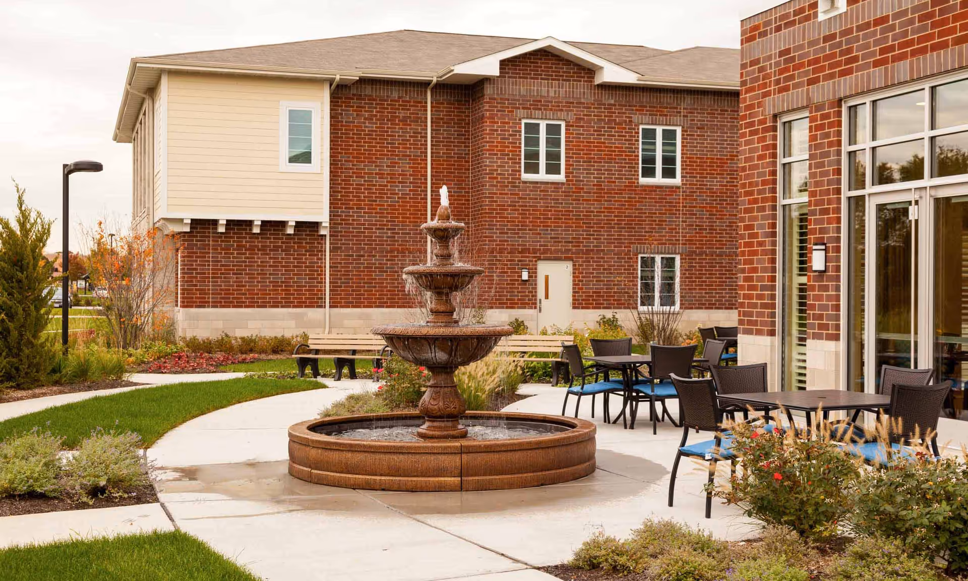 Outdoor courtyard area at HarborChase of Naperville featuring a three-tiered water fountain in the center, surrounded by a curved concrete walkway, garden beds with shrubs and flowers, benches, and patio tables with chairs. The background shows a brick building with multiple windows and a door.