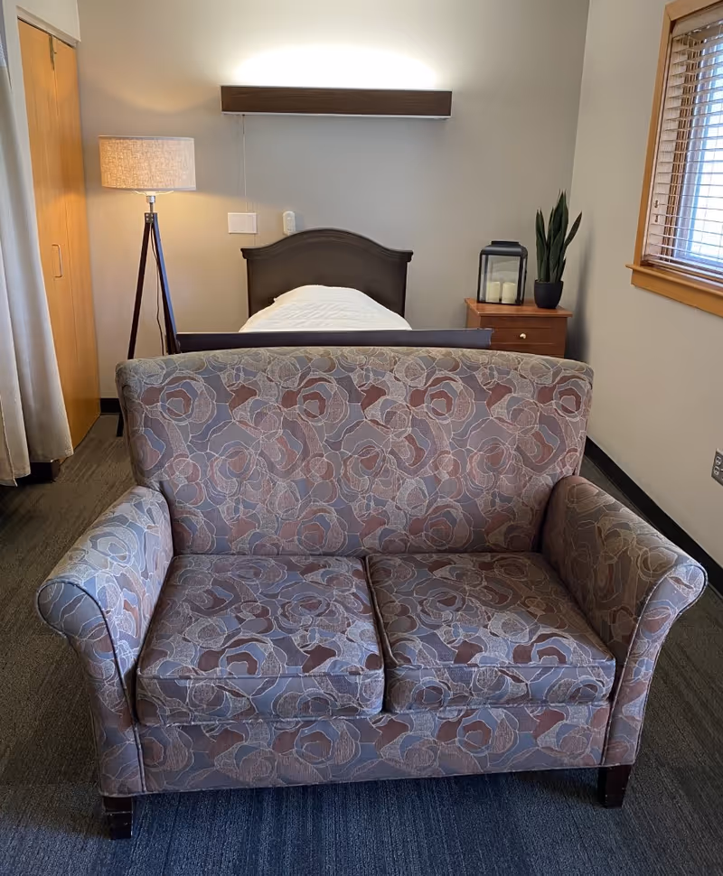 A patterned two-seat loveseat sits in front of a single bed with a bedside table, lamp, and window in a small bedroom.