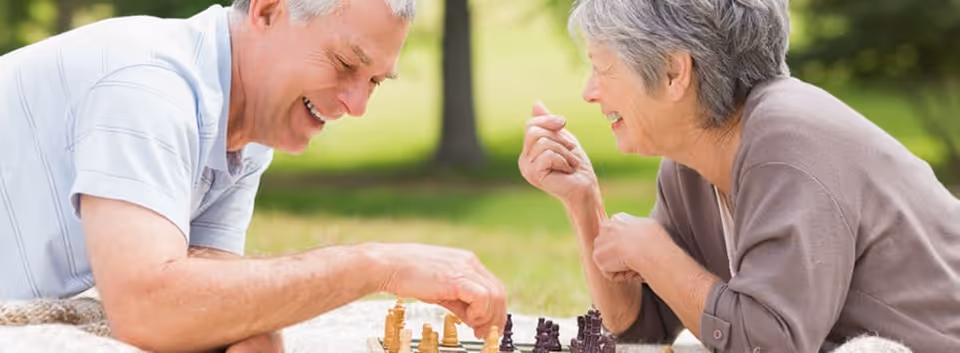 An elderly man and woman are sitting outdoors on a blanket, playing chess together and smiling, with green grass and trees in the background.