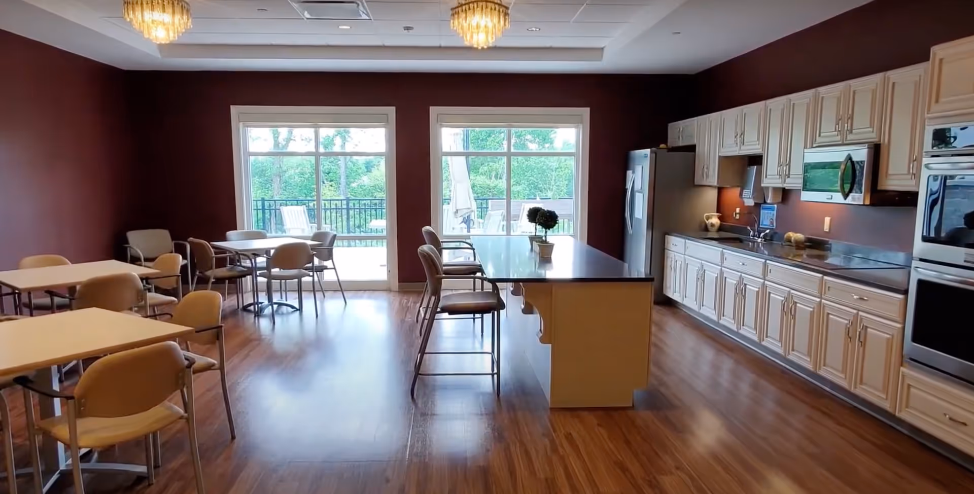 Communal dining and kitchen area with a central island, tables and chairs, and large windows overlooking an outdoor patio.