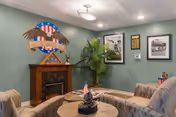 A cozy communal lounge with upholstered chairs around a small table facing a decorative mantel topped by a folded flag and patriotic emblem, a potted plant, and framed art on green walls.
