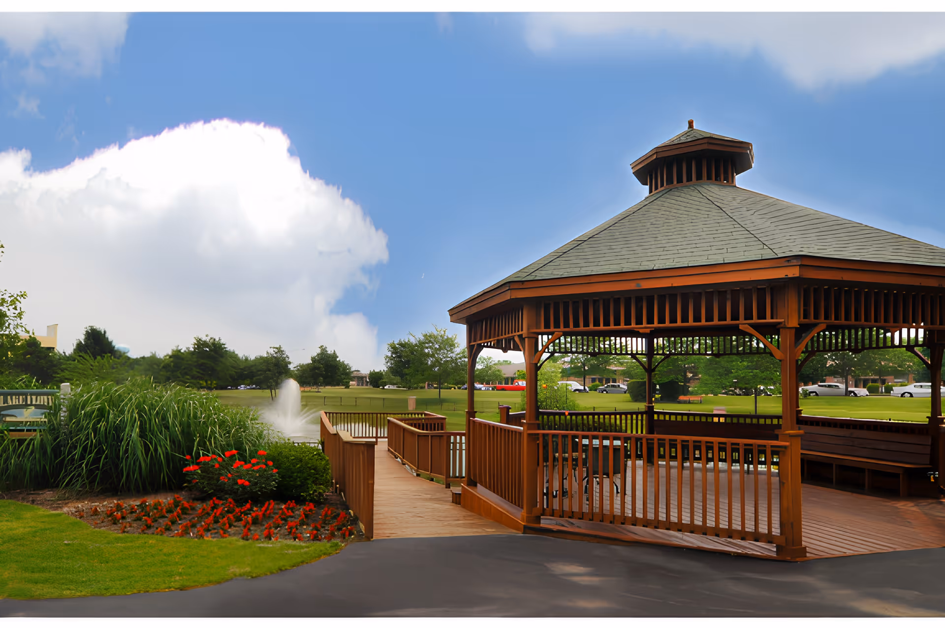 A wooden gazebo with a shingled roof situated in a landscaped outdoor area with green grass, red flowers, bushes, and a water fountain in the background under a partly cloudy blue sky.
