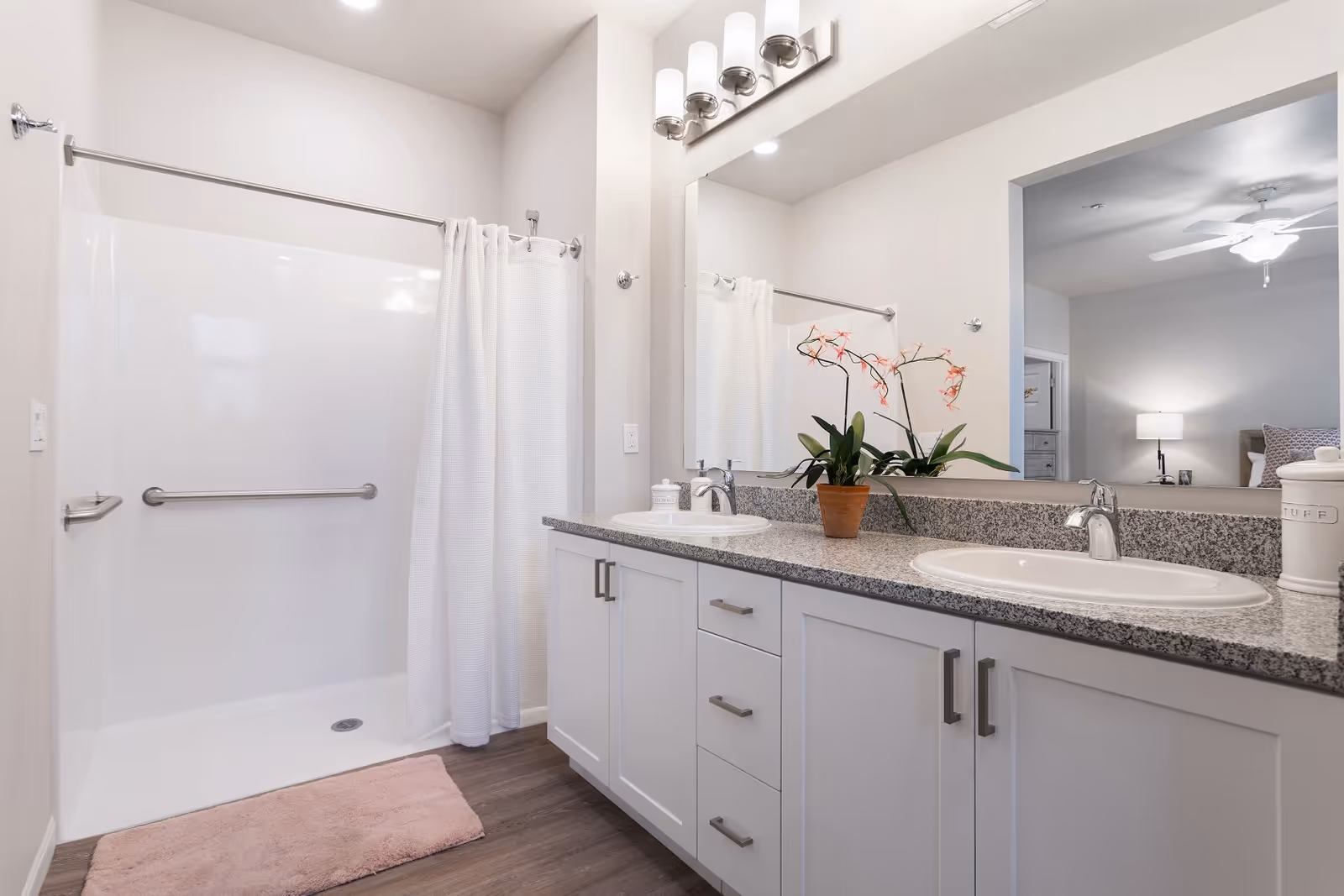 A clean and modern bathroom featuring a walk-in shower with a white curtain and grab bars, a double sink vanity with granite countertop, white cabinets, a potted orchid plant, and a large mirror reflecting a bedroom with a ceiling fan and lamp.