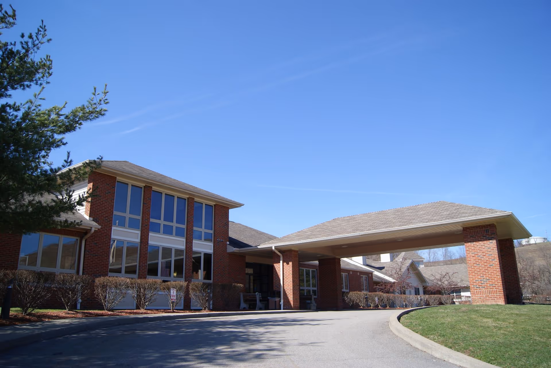 Exterior view of a brick senior living facility building with large windows and a covered driveway entrance under a clear blue sky. There are trimmed bushes along the building and a tree on the left side.
