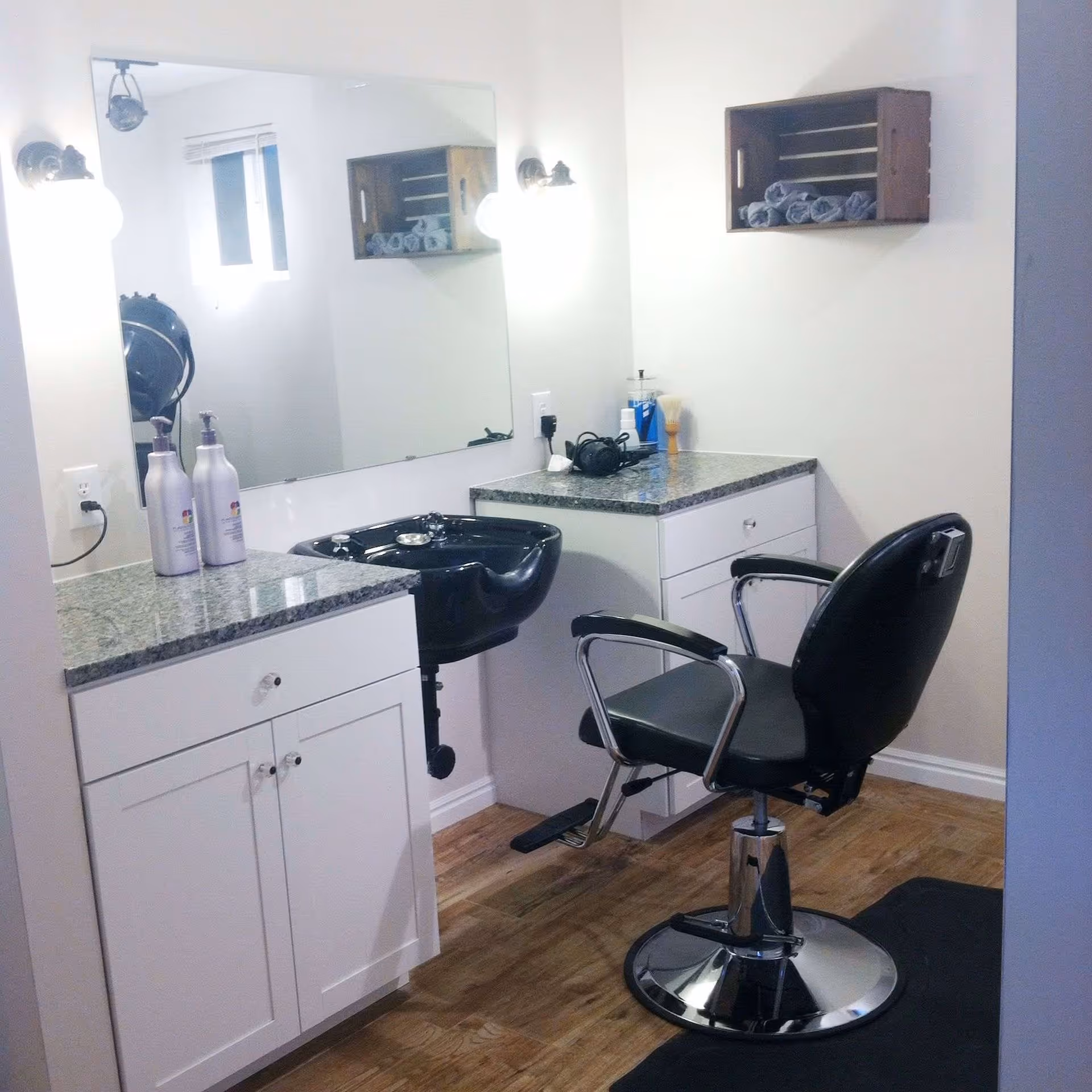 Interior view of a small salon area with a black salon chair in front of a black hair washing sink. There are white cabinets with granite countertops on either side, a large mirror on the wall, two wall-mounted light fixtures, and a wooden shelf holding rolled towels.