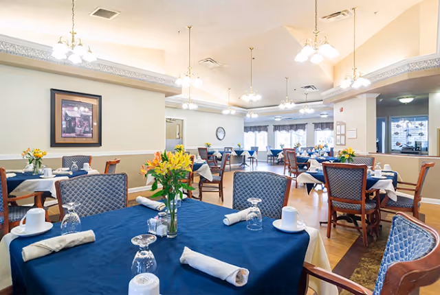 Spacious dining room with tables covered in blue tablecloths set with napkins, glassware, and flower centerpieces under hanging chandeliers.