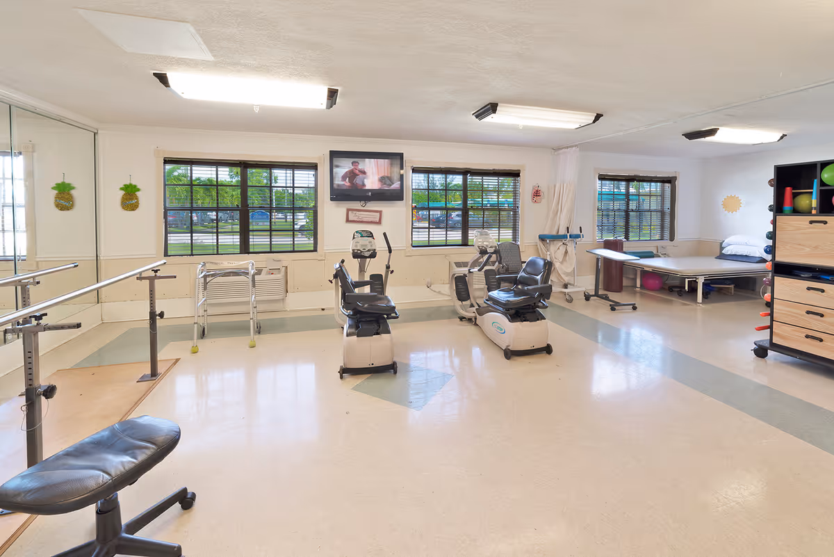 A rehabilitation room with exercise equipment including two stationary recumbent bikes, parallel bars for walking support, a padded treatment table, and various therapy balls and equipment on shelves. The room has large windows with blinds, fluorescent ceiling lights, and a TV mounted on the wall.