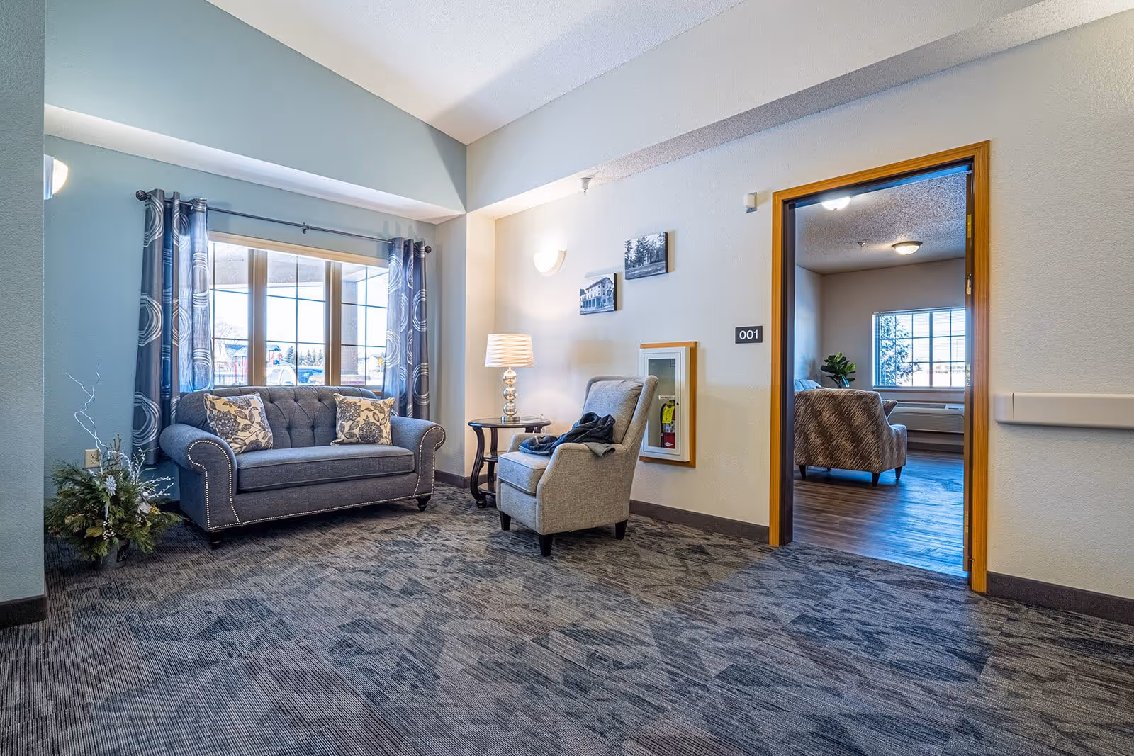 A cozy sitting area in a senior living facility with a blue loveseat adorned with two floral pillows, a gray armchair, a small round side table with a lamp, and a large window with patterned curtains letting in natural light. The room has carpeted flooring and light-colored walls with two framed pictures. An open doorway leads to another room with wooden flooring and additional seating.