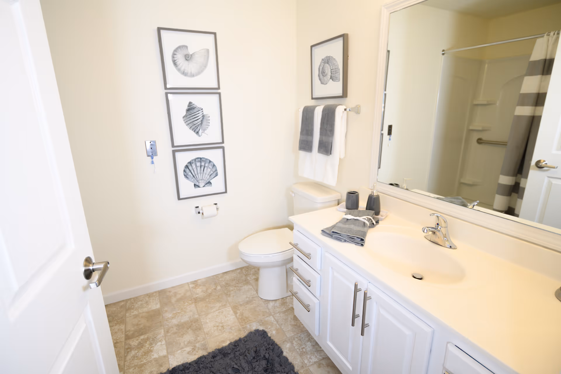 Bright modern bathroom with a white vanity and sink, toilet, mirror, shower with striped curtain, and three framed shell prints on the wall.