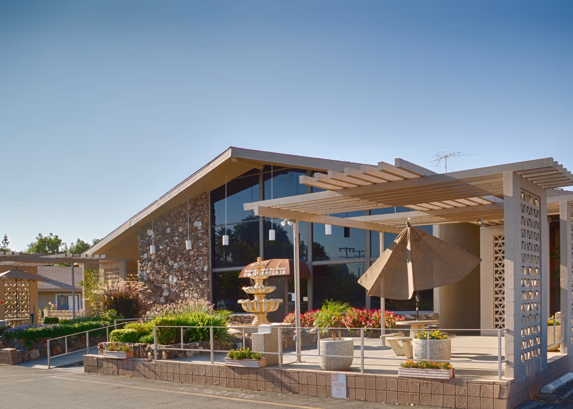 Front exterior of a stone-faced building with large windows, a pergola-covered patio, fountain, umbrella, and landscaped planters.