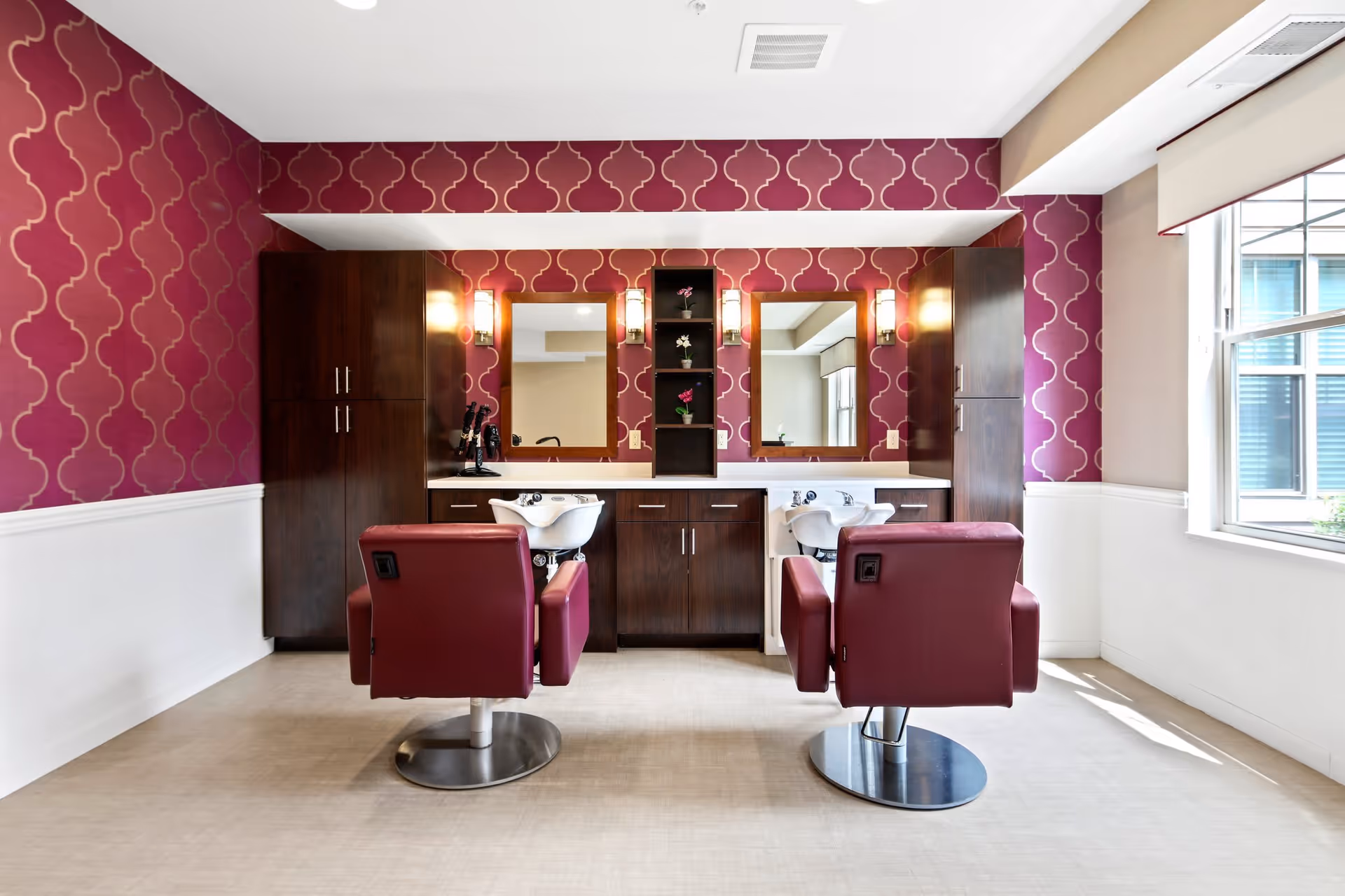 A stylish salon area with two red salon chairs facing two white wash basins and mirrors mounted on a wall with purple patterned wallpaper. There are dark wood cabinets on either side and a window on the right letting in natural light.