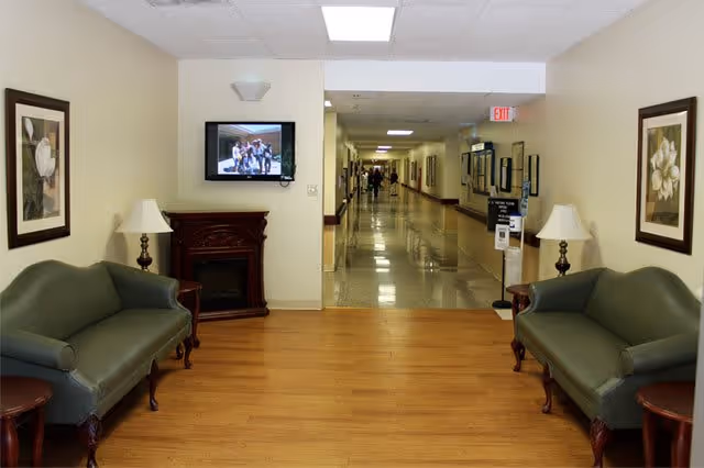 A sitting area in a senior living facility with two green upholstered sofas facing each other, two table lamps on wooden side tables, framed floral artwork on the walls, a wall-mounted television above a decorative fireplace, and a hallway leading further into the building.
