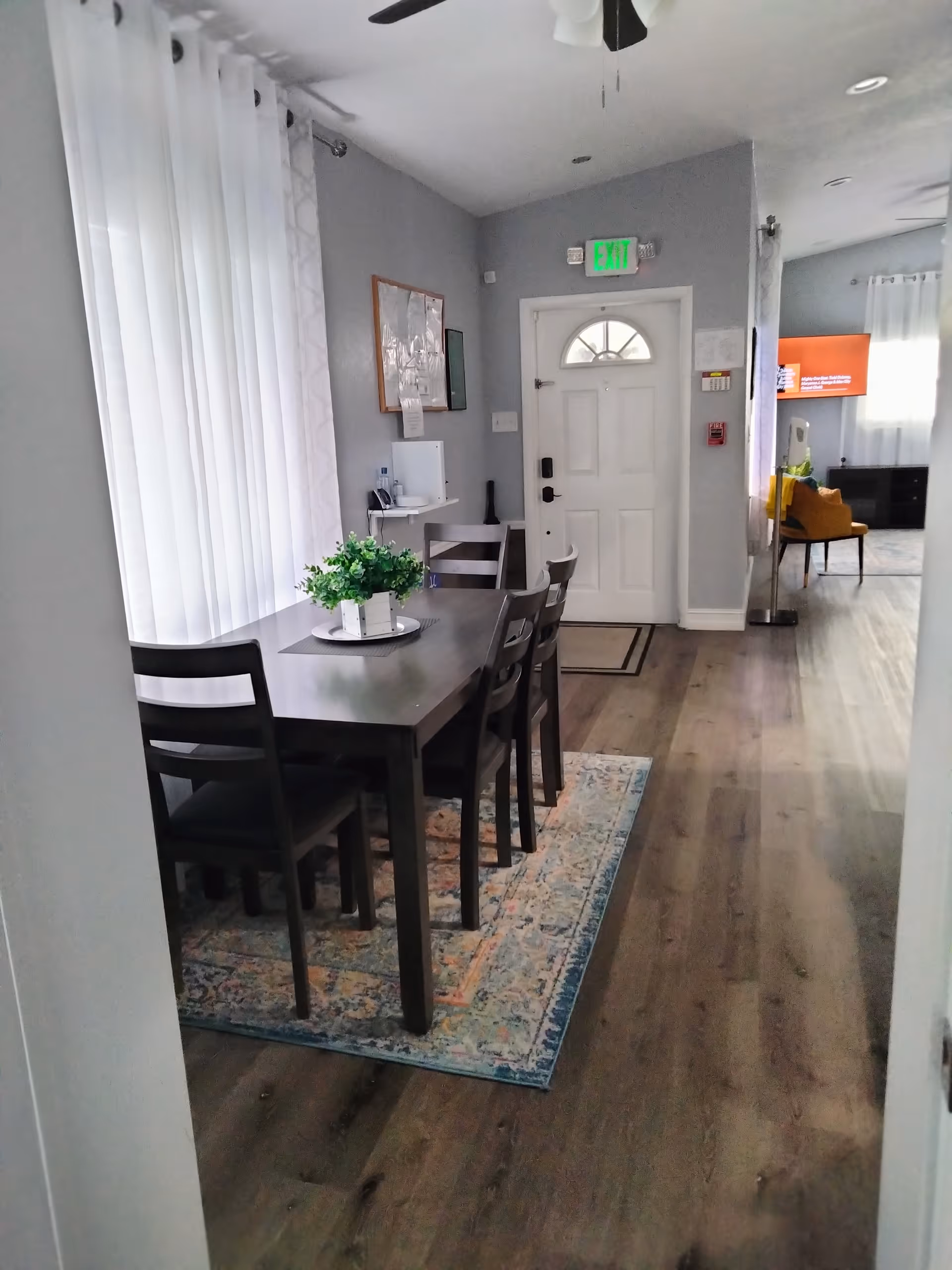 Interior view of a dining area in an assisted living facility with a dark wooden table and four chairs on a patterned rug. A small green plant centerpiece is on the table. White curtains cover the windows on the left, and a white door with an exit sign above it is visible in the background. The floor is wooden, and part of a living room with a TV and orange chair is visible further inside.