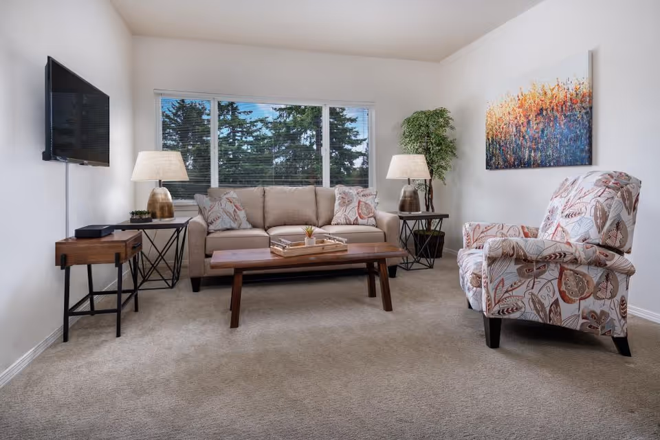 Bright living room with a beige sofa, patterned armchair, wooden coffee table, side tables with lamps, wall-mounted TV and a large window.