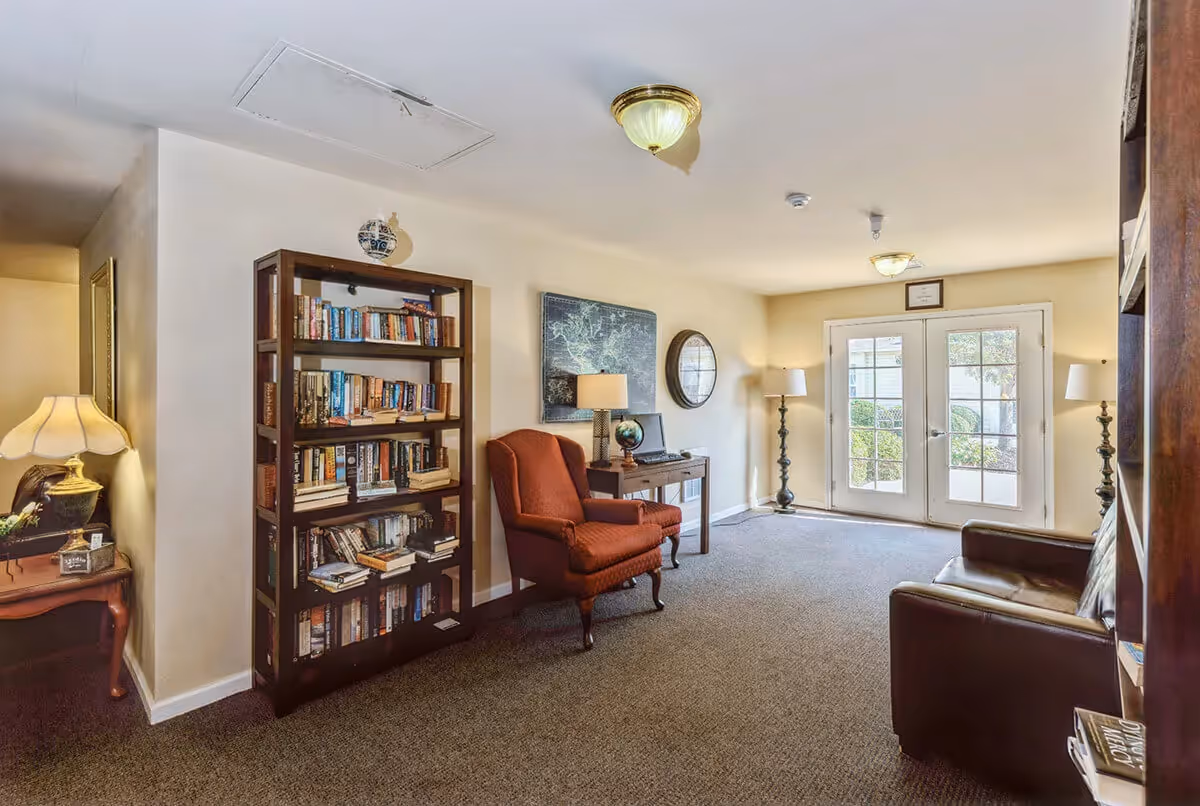 Sunlit sitting room with bookshelves, armchairs, lamps and double glass doors leading outside.