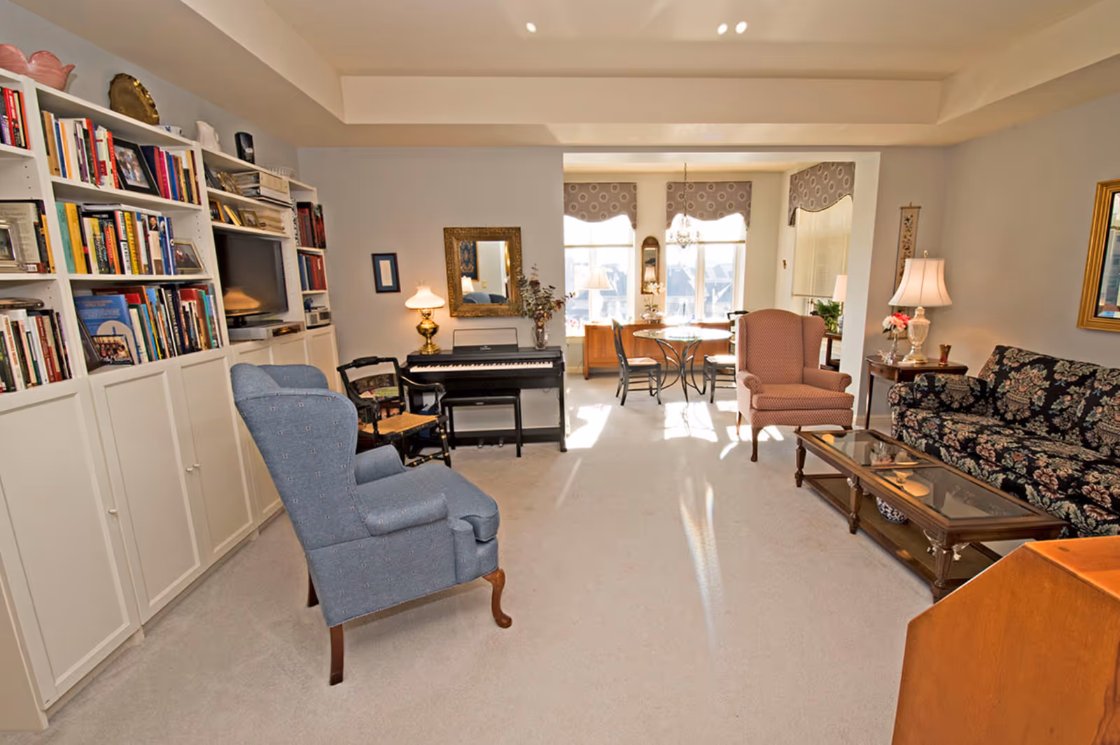 A cozy living room featuring a blue armchair, floral couch, and a dining area with a table. There is a bookshelf filled with books, a piano, and decorative elements like a lamp and a mirror.