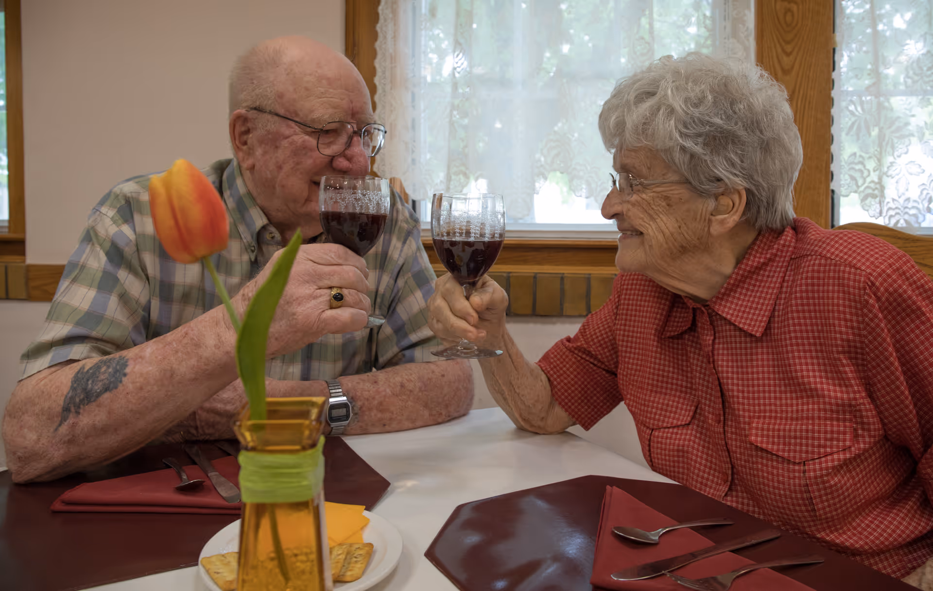 An elderly man and woman sitting at a dining table in a well-lit room, smiling and clinking glasses filled with a dark beverage. The table is set with maroon placemats, silverware, and a small vase with an orange tulip. The background shows a window with lace curtains.