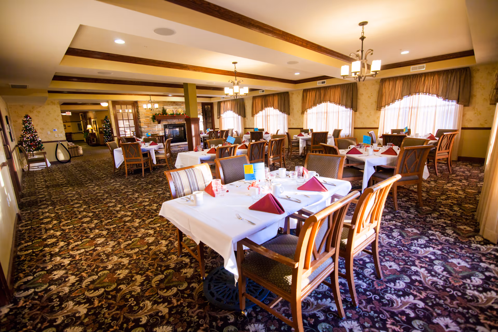 A spacious dining room with multiple tables covered in white tablecloths, each set with burgundy napkins, cups, and utensils. The room features patterned carpet, large windows with curtains, chandeliers hanging from the ceiling, and a fireplace in the background. There are Christmas trees decorated with lights near the entrance.
