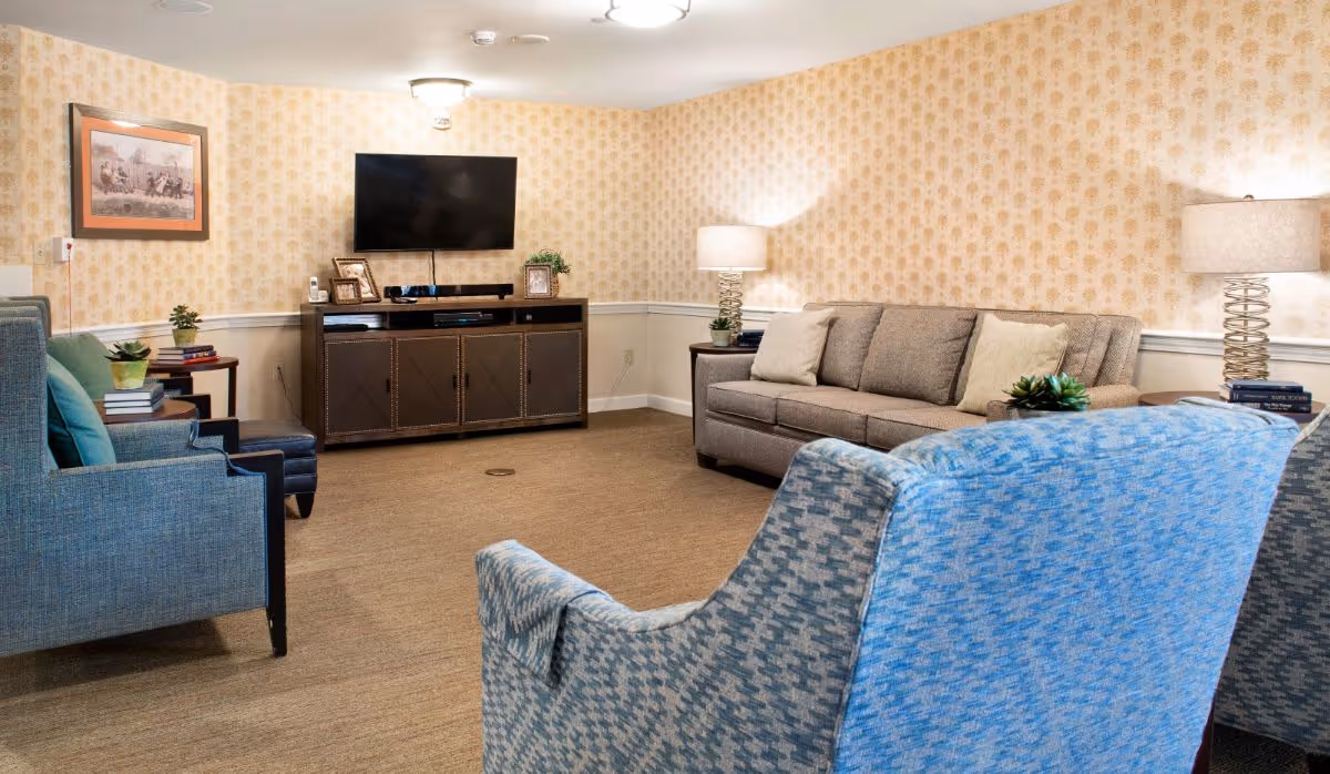 Communal living room with a sofa, several upholstered chairs, a wall-mounted TV above a console, lamps, and patterned wallpaper.