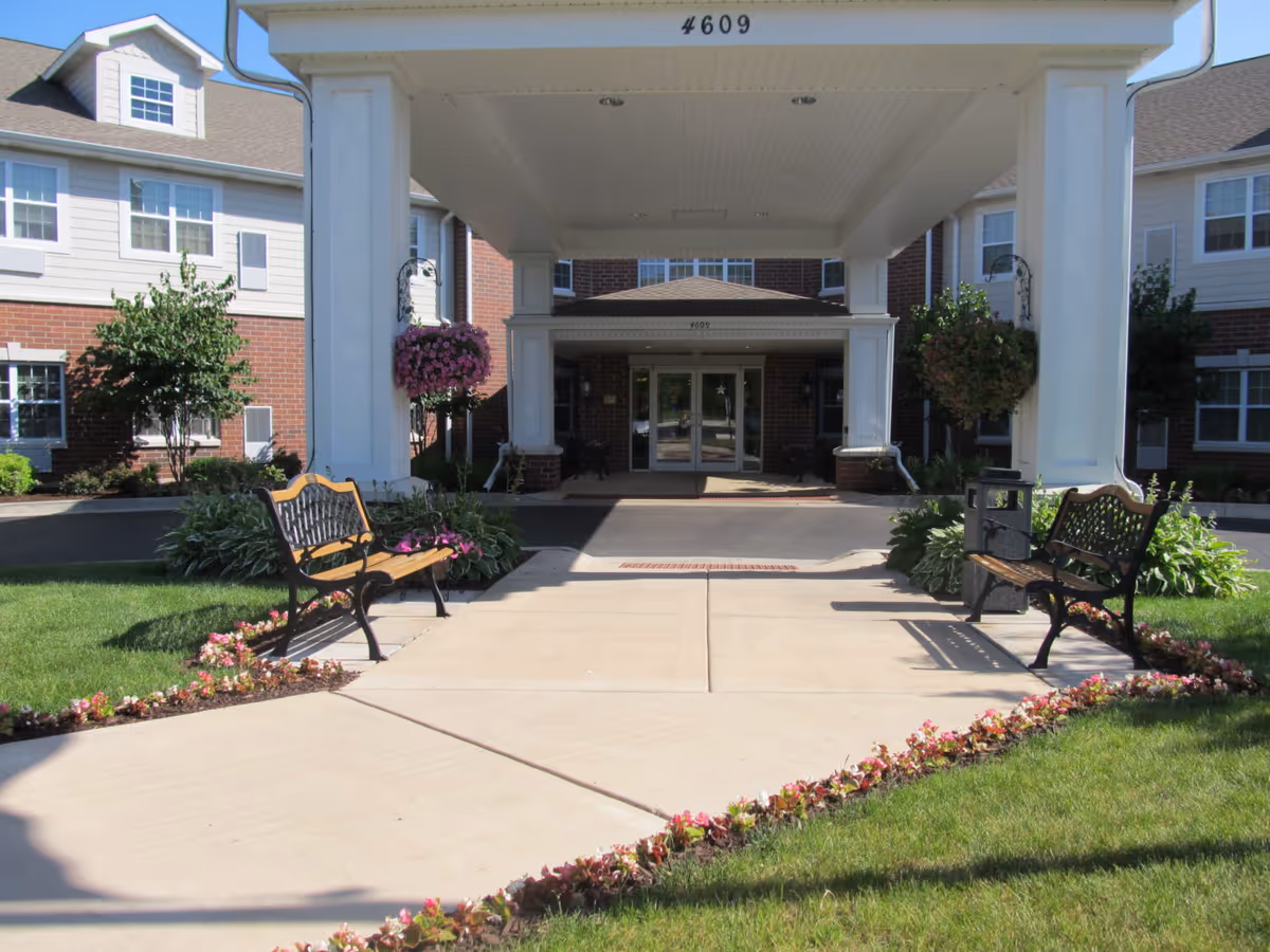 Covered entrance of a senior living building with benches, flowerbeds, and the address number 4609 above the canopy.
