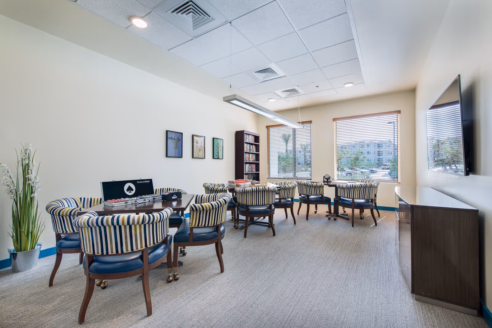 A bright and spacious common room with several tables and striped cushioned chairs arranged for group activities. One table has a poker set with chips and cards, and another table has board games. There is a tall bookshelf filled with books against the far wall, three framed pictures on the wall, large windows with blinds letting in natural light, a flat-screen TV mounted on the right wall, and a cabinet below the TV. A tall plant in a metal pot is placed in the corner.
