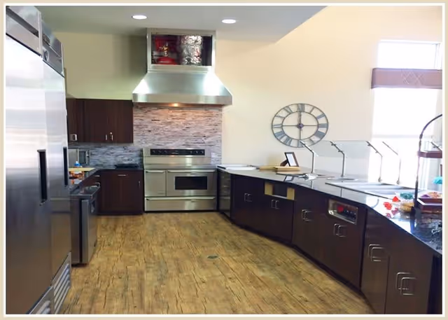 Large modern kitchen/serving area with stainless steel appliances, dark cabinets, wood-look floor, and a buffet counter under a wall clock.