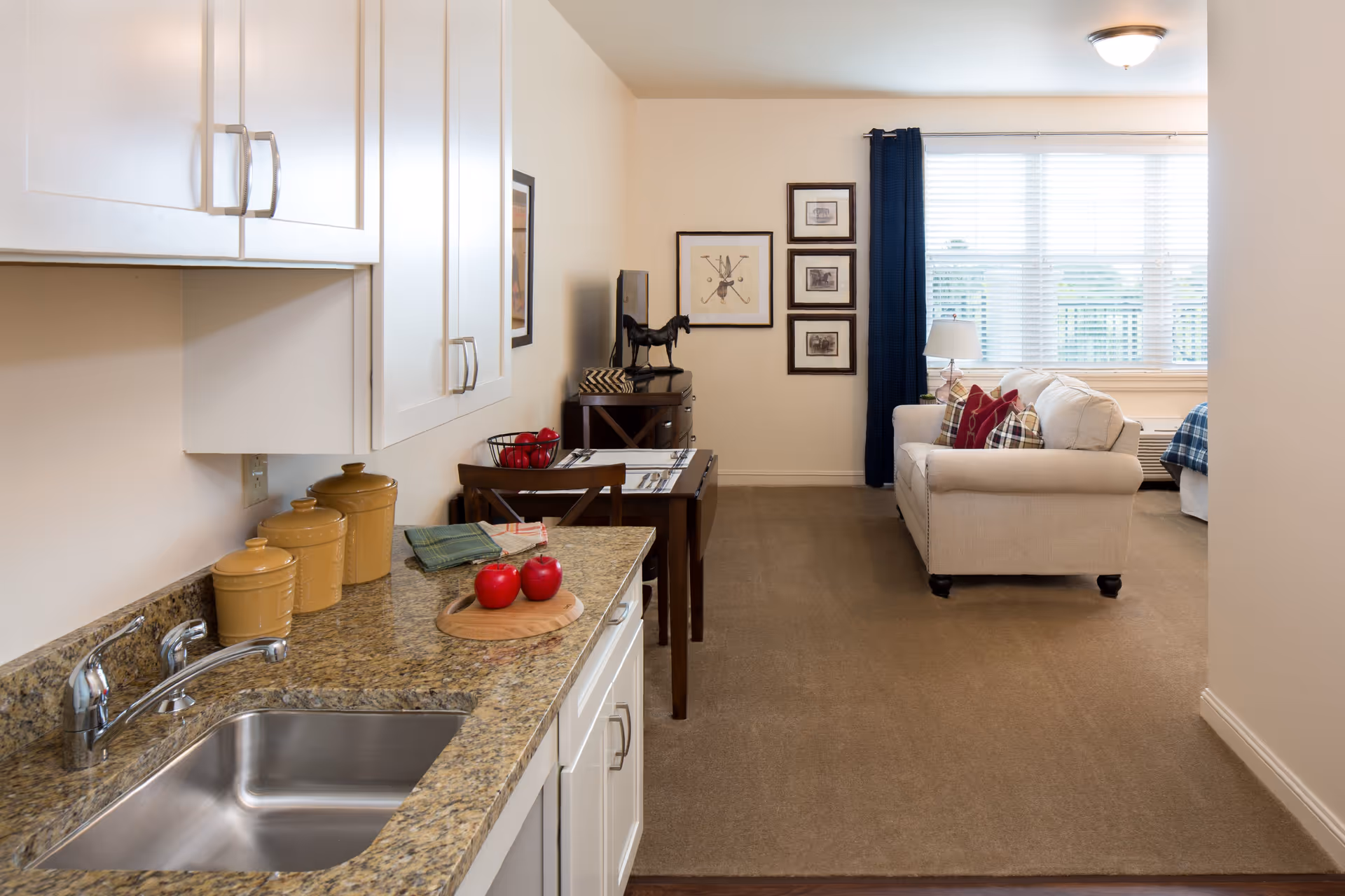 Interior view of a senior living facility apartment showing a small kitchen area with a granite countertop, stainless steel sink, and white cabinets. On the counter are three ceramic canisters and a wooden cutting board with three red apples. Beyond the kitchen is a dining table set for two with a bowl of red apples, a TV on a stand, and a beige sofa with colorful pillows in front of a large window with white blinds and dark blue curtains. The room is carpeted and well-lit with a ceiling light.