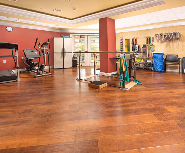 Spacious senior fitness/activity room with exercise machines, parallel bars, therapy equipment, and wood-style flooring.