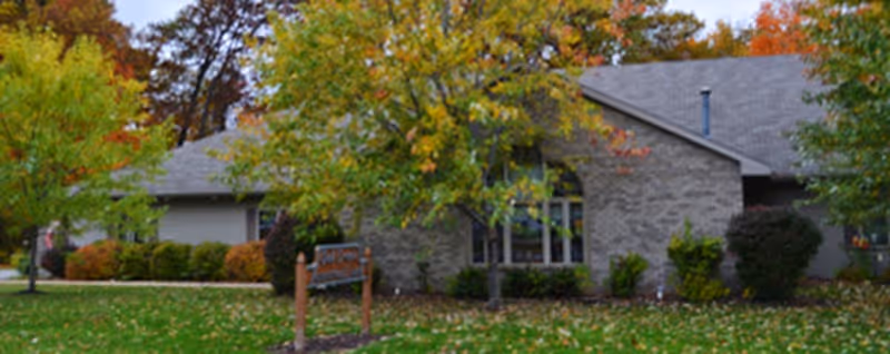 Front exterior of a single-story brick senior living building surrounded by trees with autumn foliage and a small wooden sign on the lawn.