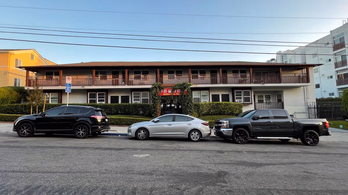 Two-story residential building with a covered upper balcony and three cars parked on the street in front.