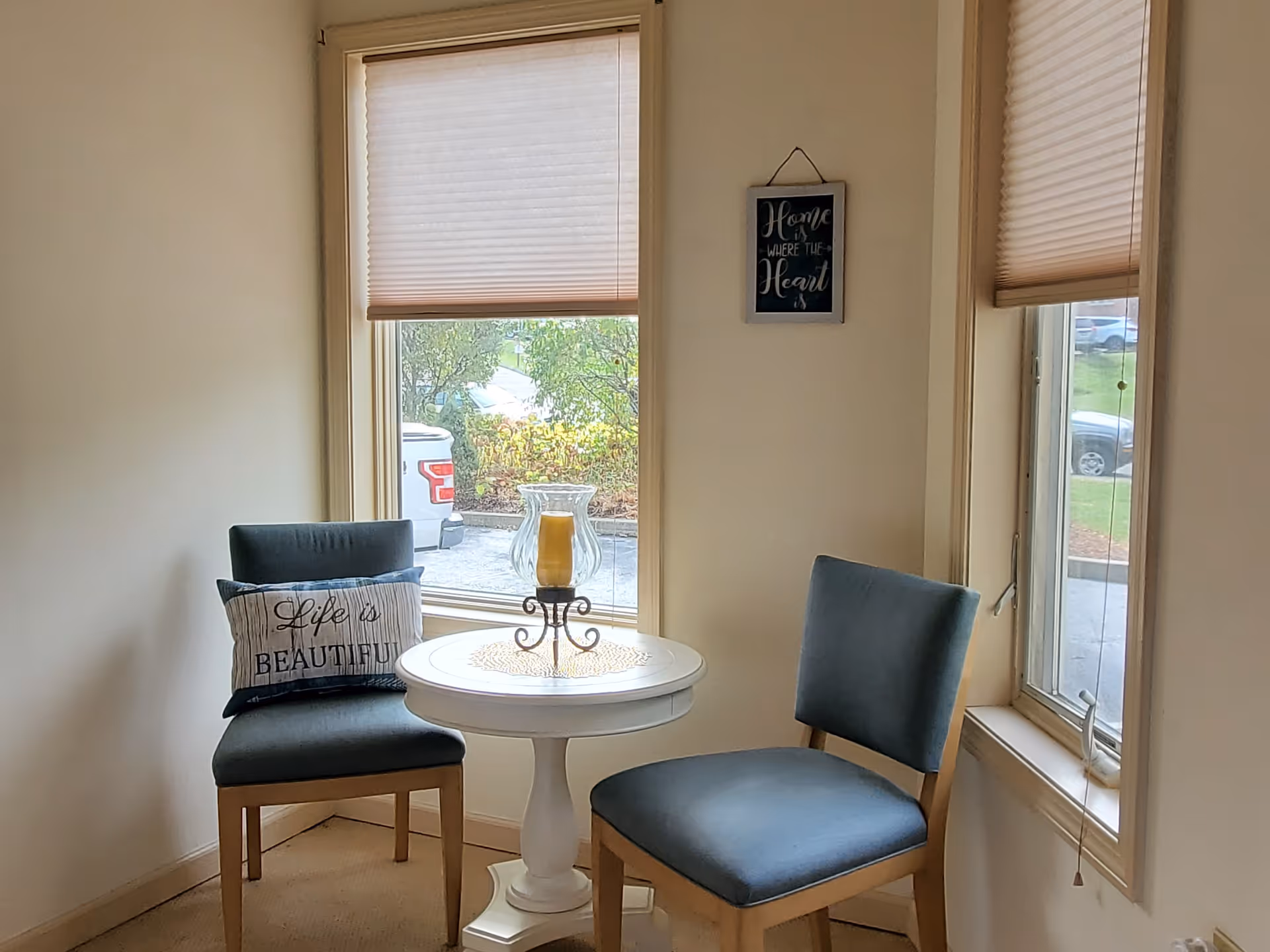 Two upholstered chairs flanking a small white round table with a decorative candle by two windows.