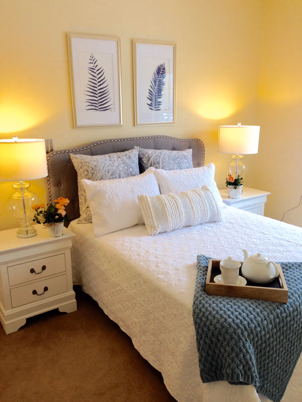 A neatly made bed with white and patterned pillows and a textured white bedspread. A blue knitted throw blanket is draped at the foot of the bed with a wooden tray holding a white teapot and cup. Two white nightstands flank the bed, each with a glass lamp and a small flower arrangement. Above the bed are two framed botanical prints of fern leaves on a light yellow wall.