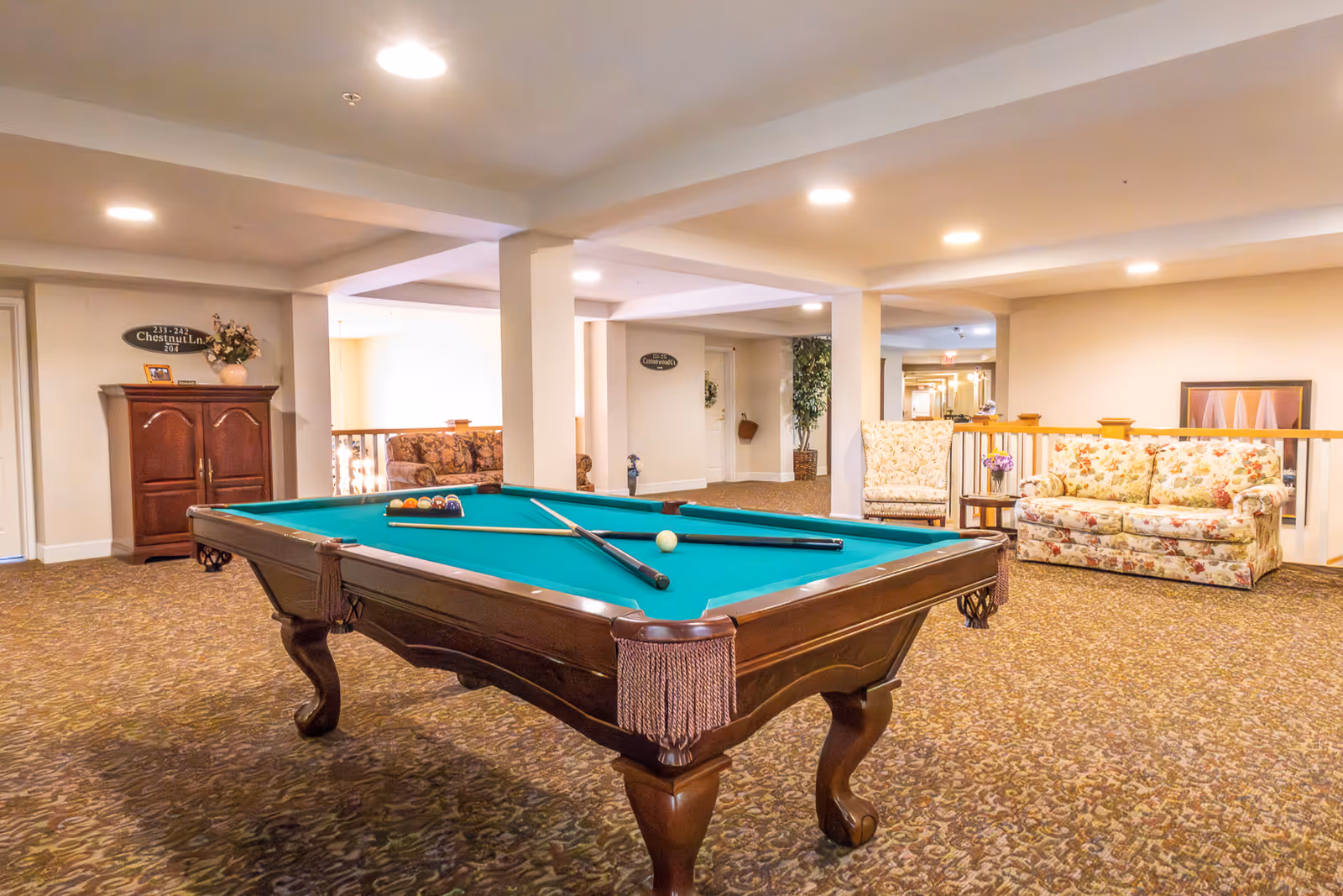 A spacious indoor recreational area featuring a pool table with cues and balls set up for a game. The room has carpeted flooring and is furnished with floral-patterned sofas and armchairs. There are decorative plants and framed artwork on the walls, with soft overhead lighting creating a warm and inviting atmosphere.