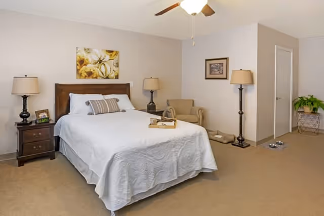 A bedroom with a neatly made bed featuring a white quilt and pillows, two bedside tables each with a lamp, a beige armchair in the corner, a framed picture on the wall, a ceiling fan with light, and a small plant on a stand near a closed door. There are also pet bowls and a pet bed on the floor.